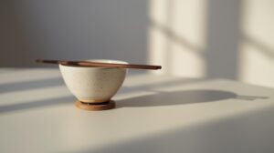 夕方のやわらかな自然光が差し込む白いテーブルに、陶器の茶碗と箸が箸置きに整えられ、食べる直前の静けさが漂う情景